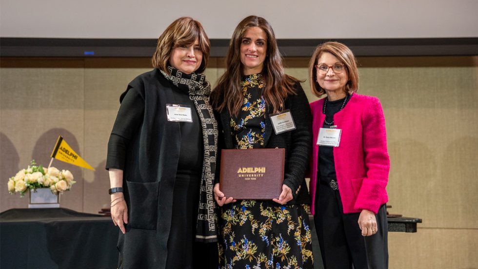 alt text: [Three women standing together facing the camera. Woman in center holds a brown award certificate case marked Adelphi University.