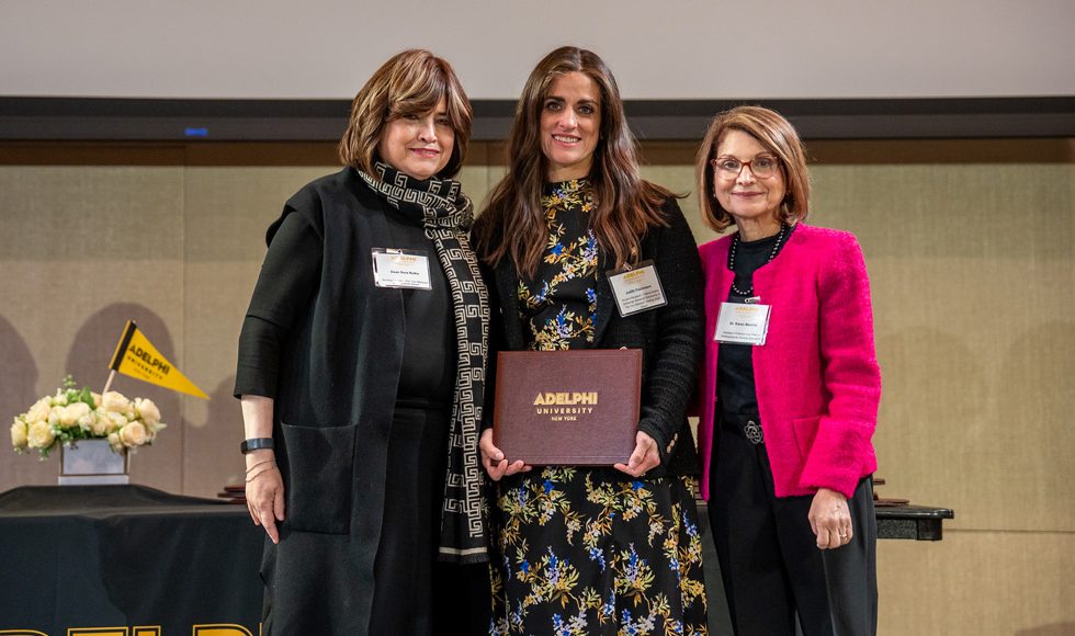  alt text: [Three women standing together facing the camera. Woman in center holds a brown award certificate case marked Adelphi University.