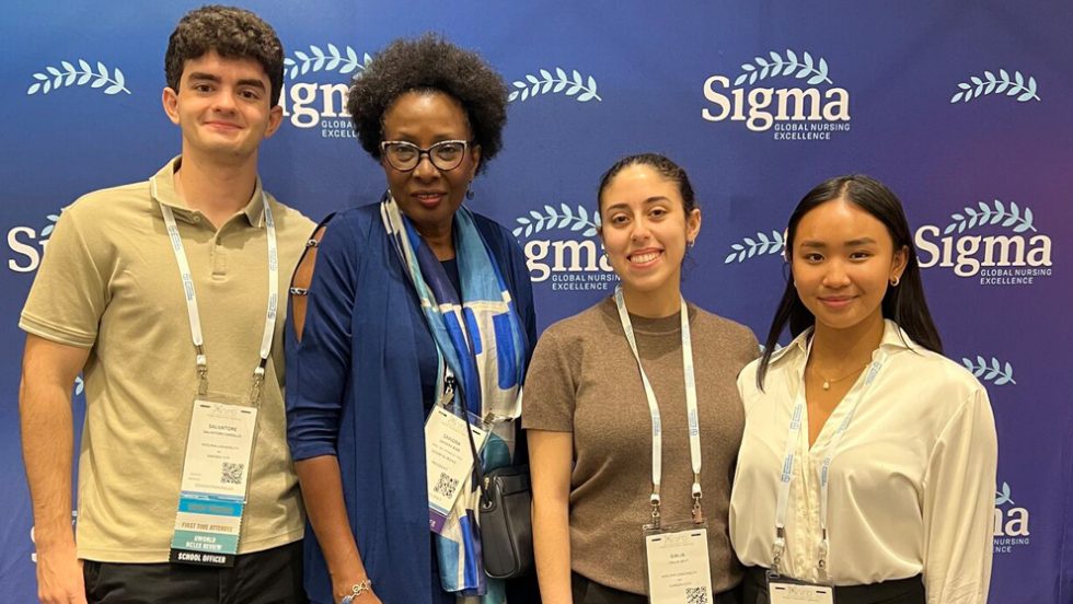 One male and three female students wearing neck lanyards stand in front of a wall labeled Sigma Global Nursing Excellence.