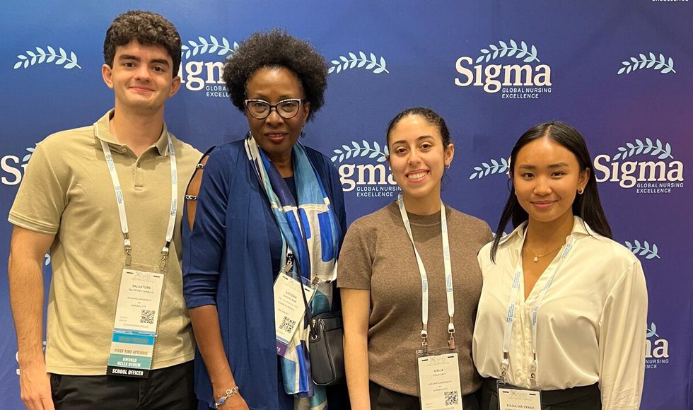 One male and three female students wearing neck lanyards stand in front of a wall labeled Sigma Global Nursing Excellence. 