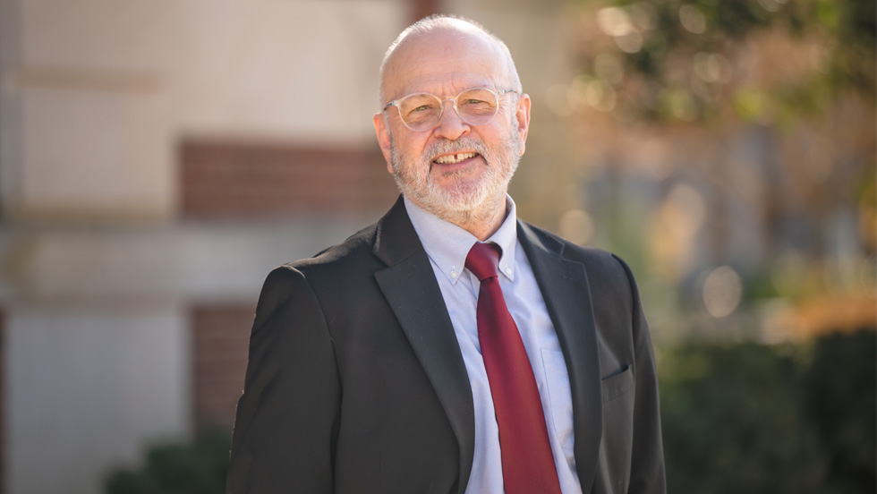 Smiling older man with a white beard and glasses, wearing a black suit jacket, light blue shirt, and red tie, standing outdoors in a sunny setting with blurred greenery and a brick building in the background.