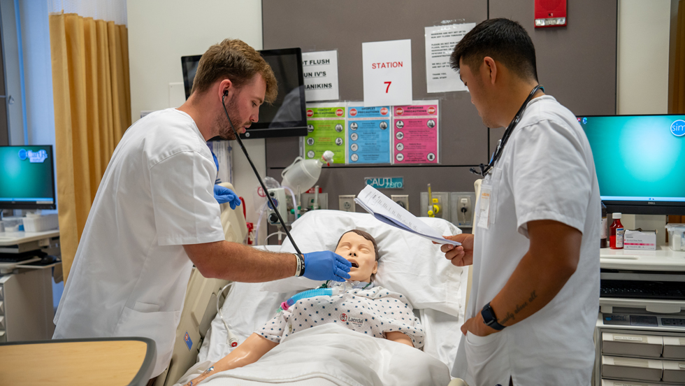 Two male nursing students in the nursing simulation lab, checking vital signs on a lifelike manikin in a hospital bed