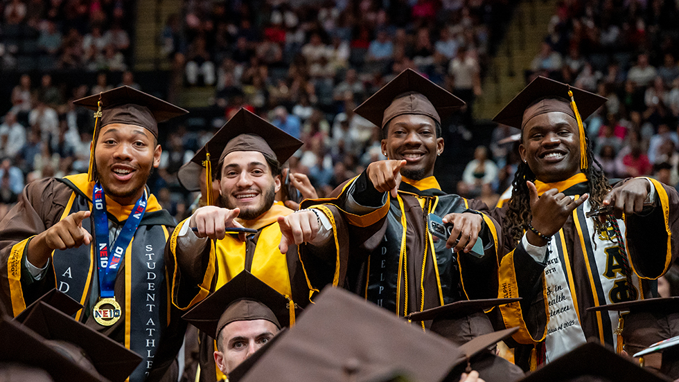 Four students smiling and posing in graduation regalia among their classmates