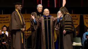 A man in graduation regalia standing with his arms up while receiving an honorary doctorate surrounded by a group of university officials