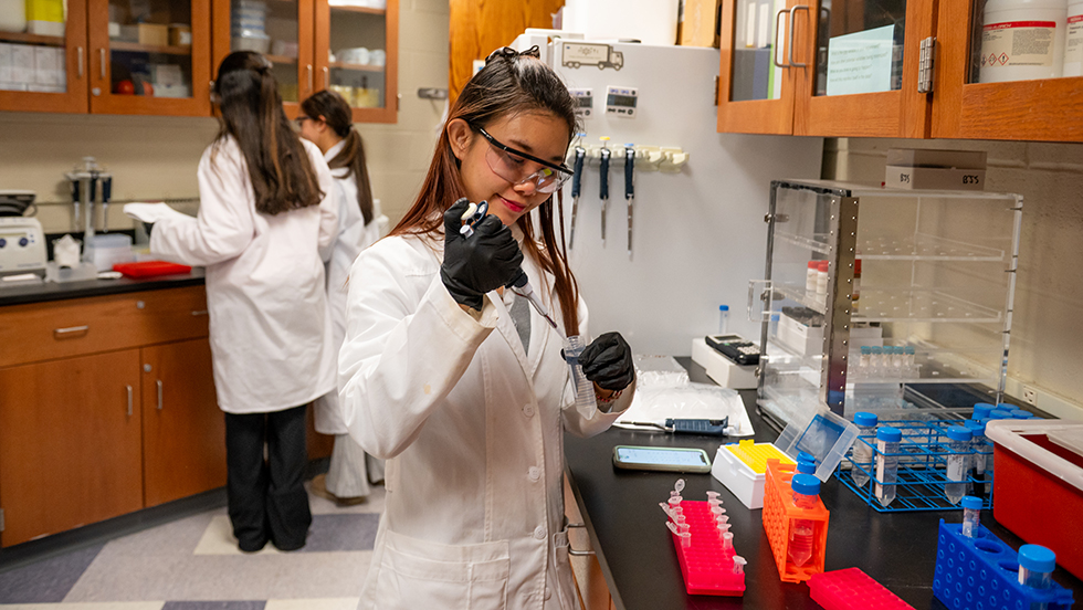 A young woman in a lab coat is focused on her work at a laboratory bench, surrounded by scientific equipment.