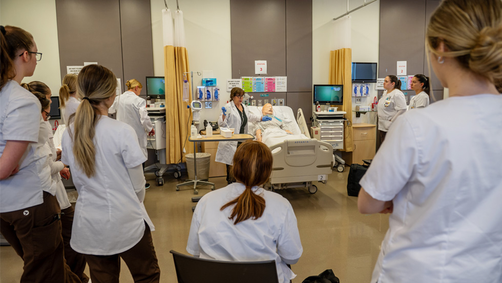A group of nursing students wearing white uniforms attentively surrounding a patient mannequin in a simulated hospital room, providing care and support.