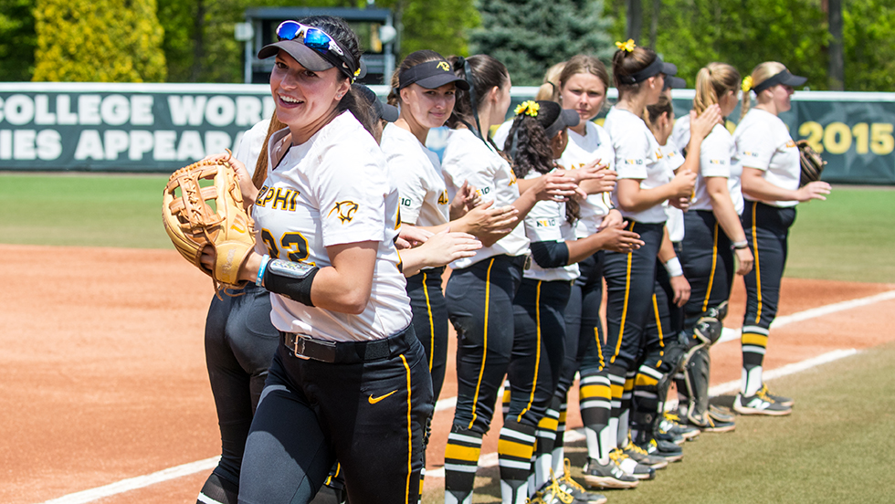 A uniformed woman softball player, posing with her team, turns to smile at the camera.