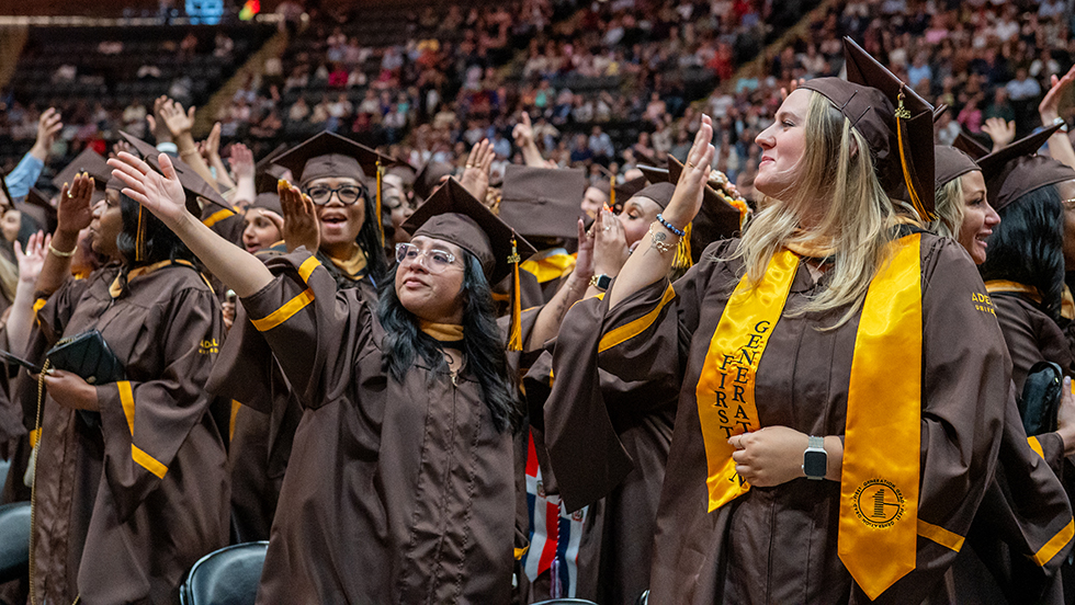 In a crowd of graduates on Commencement day, one grad blows a kiss and the other waves to family and friends.