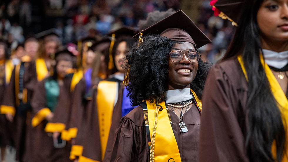 A line of students in graduation gear files into Commencement; one student smiles proudly.