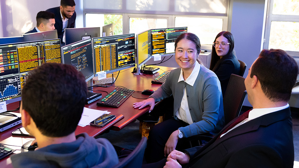 Undergraduate business students sit in front of computers with teacher looking on and smiling.