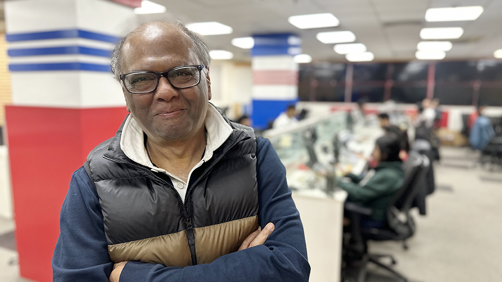 Kajaria, dressed casually, at the office with employees at work on computers at their desks in the background.