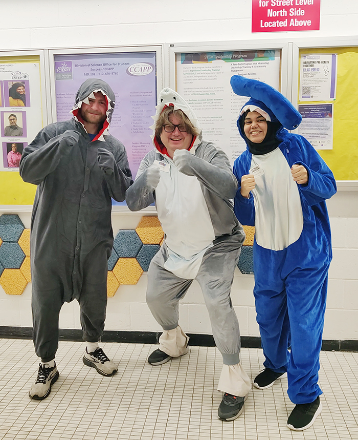 The three pose dressed in full-body shark costumes. Behind them are bulletin boards with academic postings.