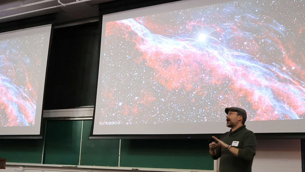Dr. Coyne stands onstage in front of a projected image of the Veil Nebula.