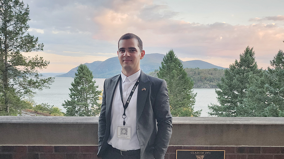 Mikautadze, wearing a gray suit with a lapel button of the U.S. and Georgia flags, stands on the Military Academy campus on a beautiful late afternoon with the Hudson River and Storm King Mountain in the background.