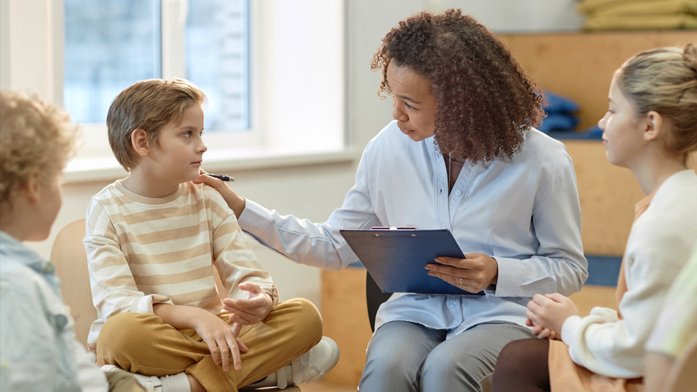 A counselor holding a clipboard talks to a young boy while sitting with a small group of children in a classroom setting.
