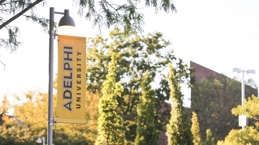 Gold and black banner flag that says Adelphi University displayed on a pole with the Garden City Campus in the background.