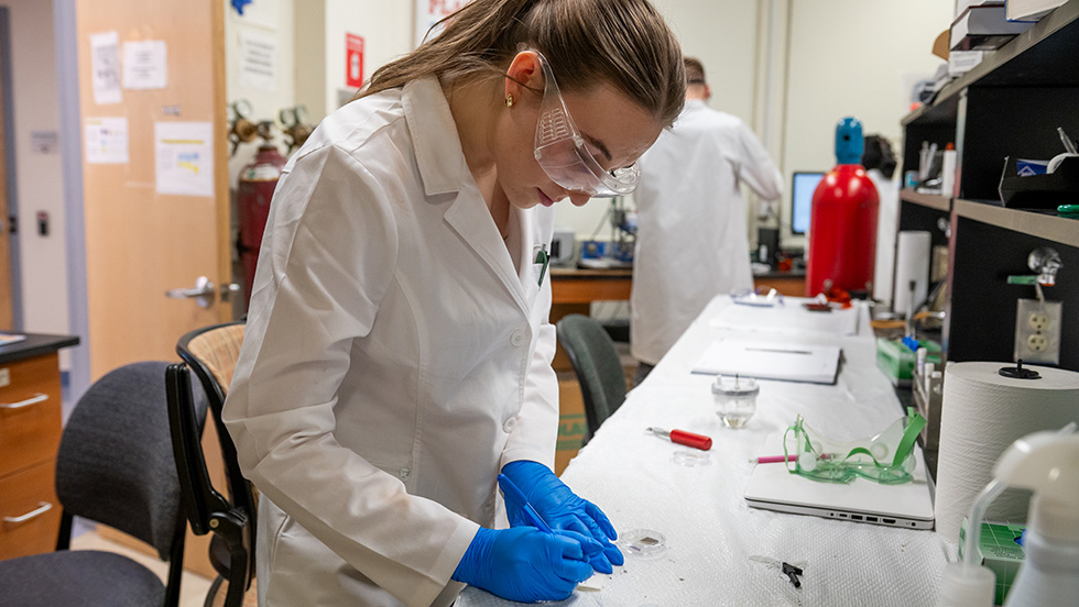 A female research in a lab coat and safety glasses performs research in an Adelphi science lab.