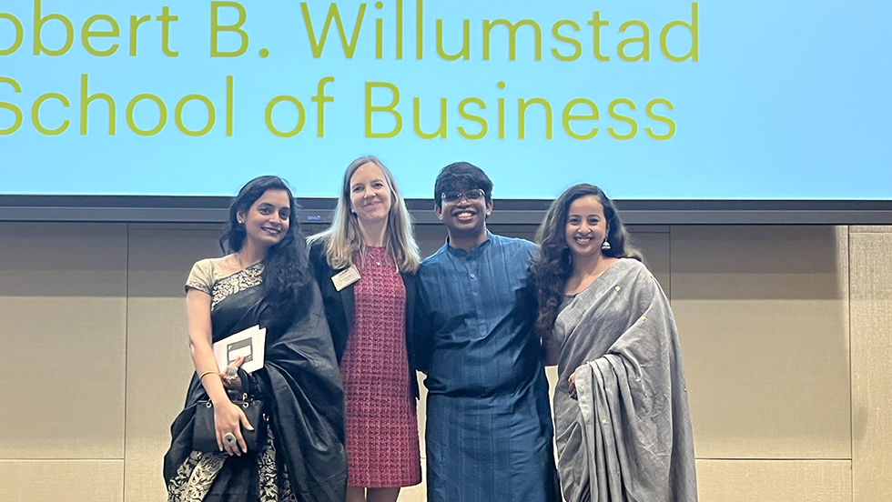 Four people smiling in front of a screen that reads “Robert B. Willumstad School of Business.”