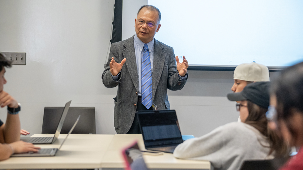 A professor speaks to students in a classroom, with laptops open on desks.