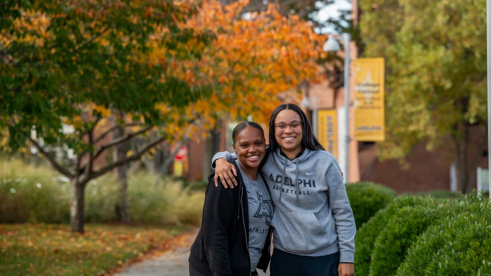 Two women standing side by side smiling, wearing Adelphi gear, on Adelphi's Garden City campus