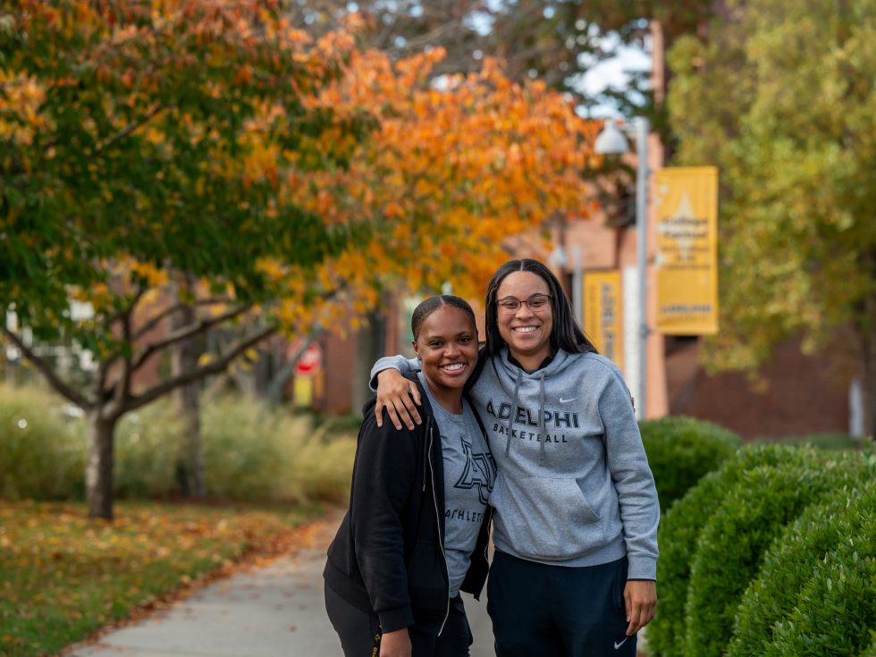 Two women standing side by side smiling, wearing Adelphi gear, on Adelphi's Garden City campus