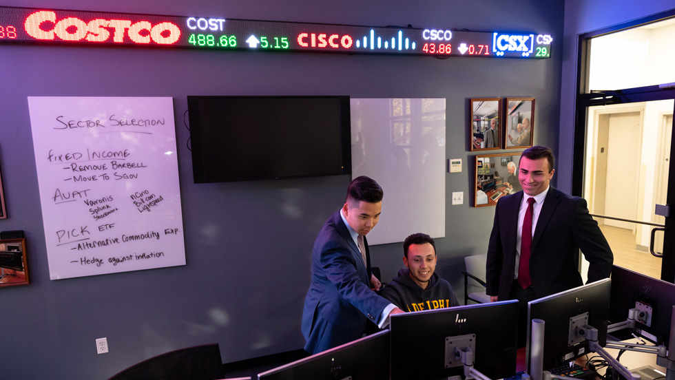 Three students gather around a computer terminal in the trading room. A digital stock ticker and a whiteboard are on the wall behind them.