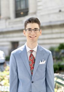 An Honors College student in a blue suit, Vincent Calvagno, stands on a city street, smiling proudly.
