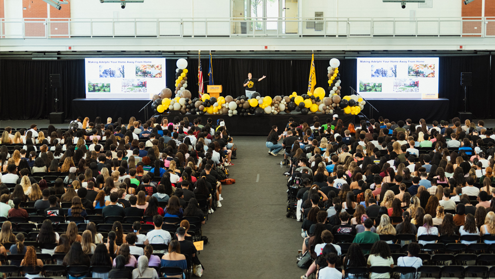 A speaker standing behind a table filled with white, brown and gold balloons addresses a large audience of new students.