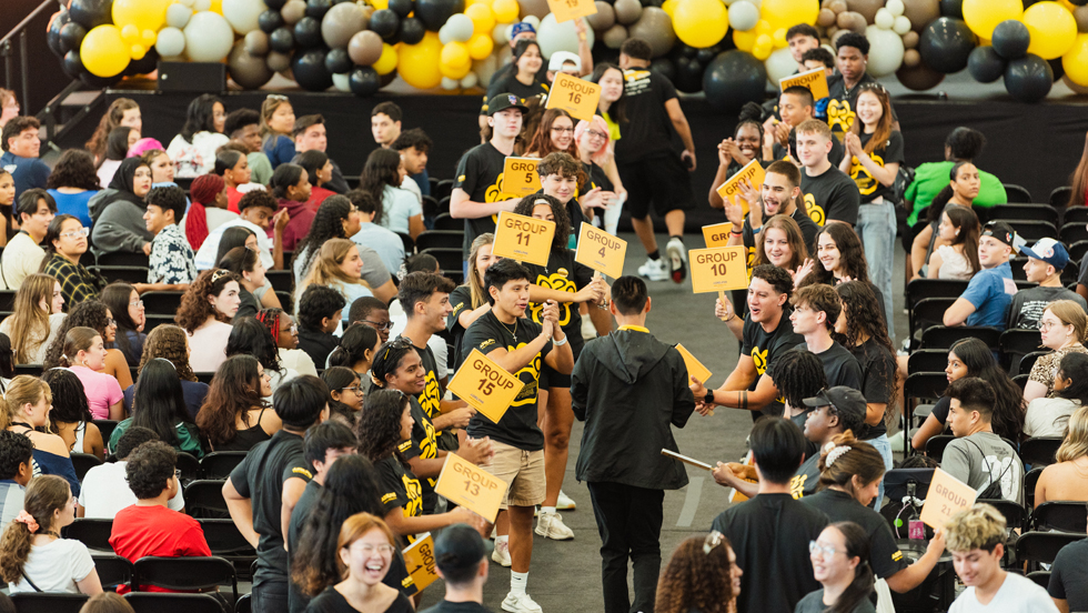 A new student walks down an aisle surrounded by students in chairs. The peer assistant leaders hold up signs identifying the different orientation groups.
