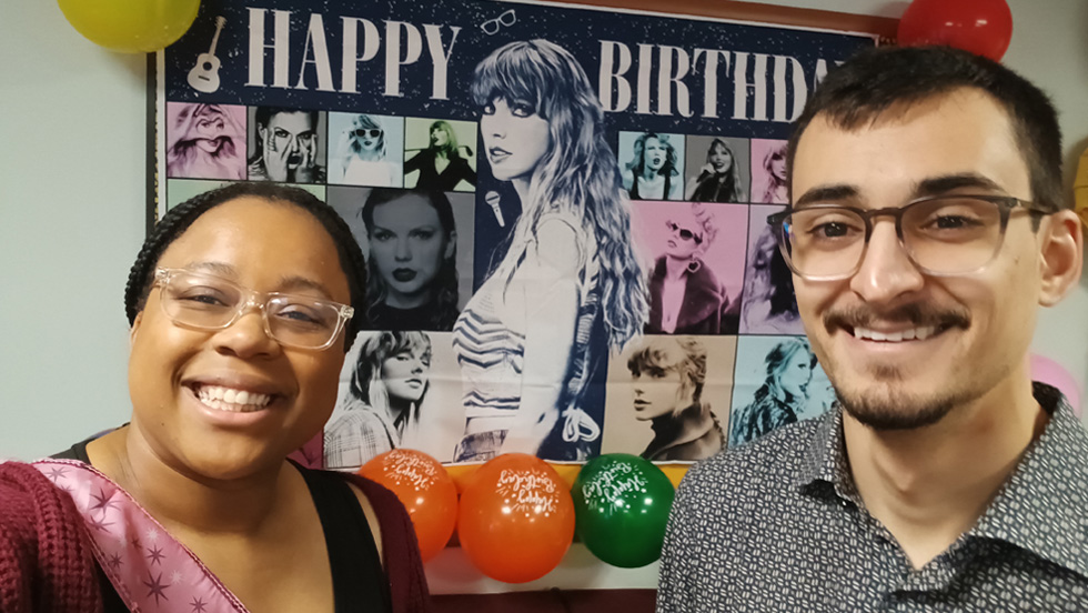 A selfie of two graduate students, a woman and a man, smiling in front of a Taylor Swift-themed birthday banner. The woman on the left, who has braided hair and is wearing glasses, is an African American with a broad smile. The man on the right, who has a beard and is wearing glasses, is a Hispanic individual with a friendly smile. Balloons are visible at the bottom of the photo.