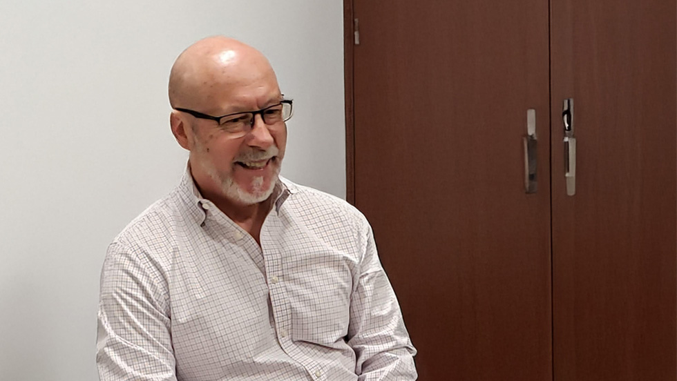 A middle-aged Caucasian man with a bald head and a gray beard is sitting and looking to the left with a smile on his face. He is wearing glasses and a light-colored, patterned button-down shirt. The background is a plain white wall and a brown cabinet or door.