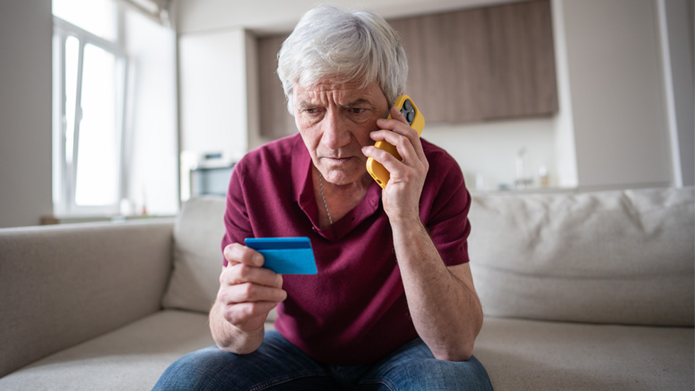 A close-up, indoor shot of a senior Caucasian man with gray hair, looking intently at a blue credit card he is holding in his left hand. He has a concerned or serious expression on his face while he talks on a yellow cell phone he is holding to his right ear. He is wearing a maroon polo shirt and blue jeans, sitting on a light-colored couch. The background is a modern living room or kitchen.