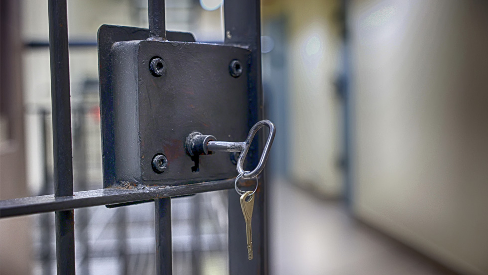 A close-up of a locked gate with a large, black lock plate and a key in the keyhole. The key, which has a ring with two smaller keys attached, is hanging from the lock. The gate is made of black metal bars and the background is out of focus, showing a blurred, institutional hallway.