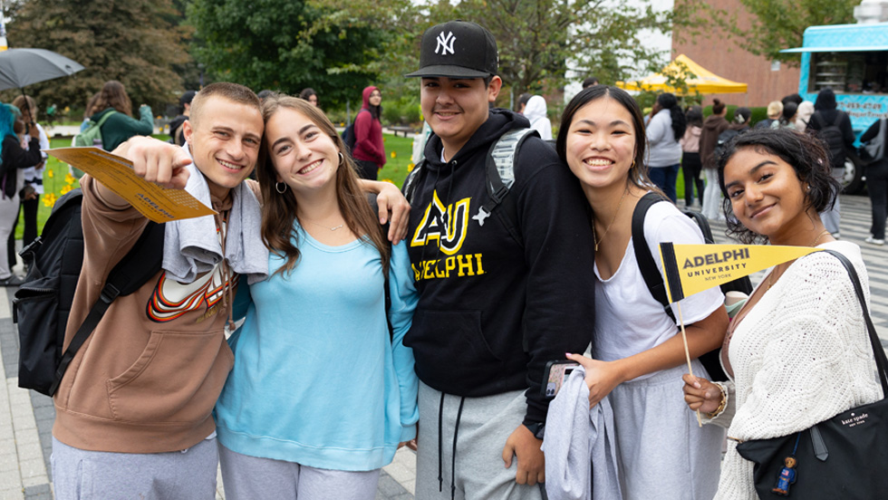 Five students, one wearing an Adelphi sweatshirt, and another holding an Adelphi pennant, pose for the camera on the University's quad.
