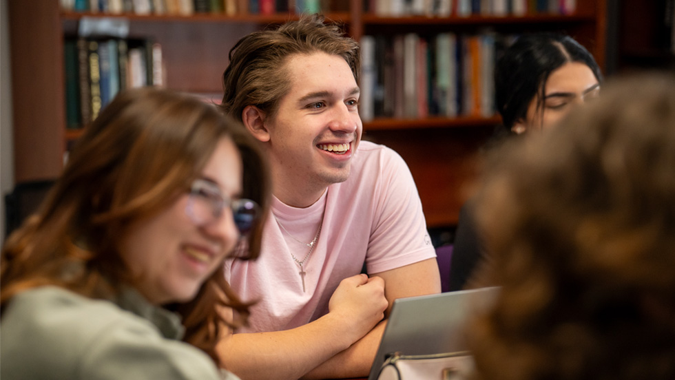 A student at a seminar table, with a student on each side of him in soft focus. Bookcases line the seminar room's walls.