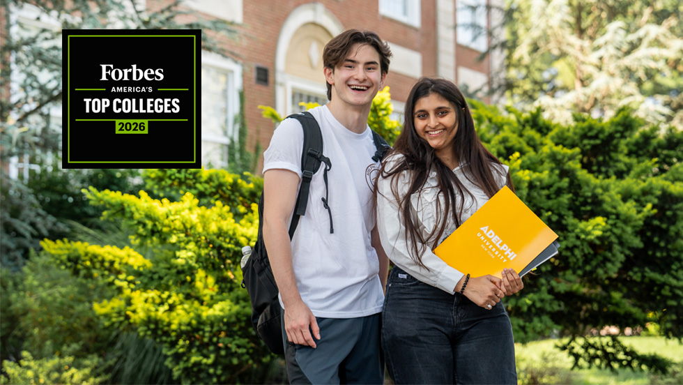 A male and female student in front of a campus building.  The woman holds an Adelphi folder.