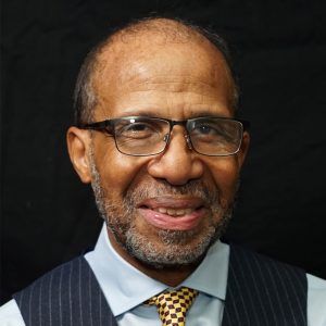 A portrait of an African American man with a bald head and a gray beard, smiling directly at the camera. He is wearing glasses, a light blue collared shirt, a dark pinstripe vest, and a checkered yellow tie. The background is a plain black wall.