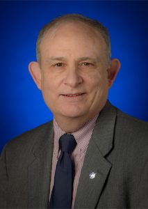 A man wearing a grey suit, striped shirt, and navy tie, with a NASA pin on his lapel, set against a blue background