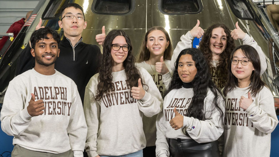A professor and group of students posing wearing Adelphi University gear giving the thumbs up.