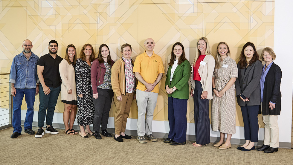 New faculty members and Adelphi leadership, all in casual attire, line up against a yellow wall for a group photo.