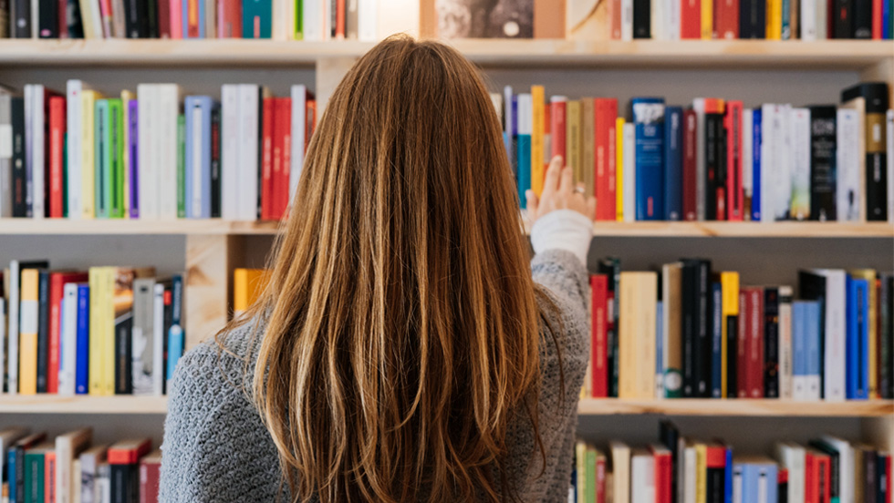 A woman with long brown hair, seen from behind, is reaching out toward a bookshelf filled with colorful books. She is wearing a gray sweater and appears to be selecting a book in a library or bookstore setting.