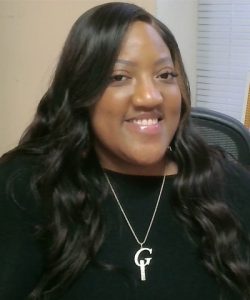 A young Black woman with long, wavy dark hair is smiling and looking directly at the camera. She's wearing a black top and a silver necklace with a 'G' pendant that has a small key design. She is seated in what appears to be an office setting, with a framed print on a desk behind her that says "TRUST THE process" in white and gold lettering over an abstract, colorful landscape.