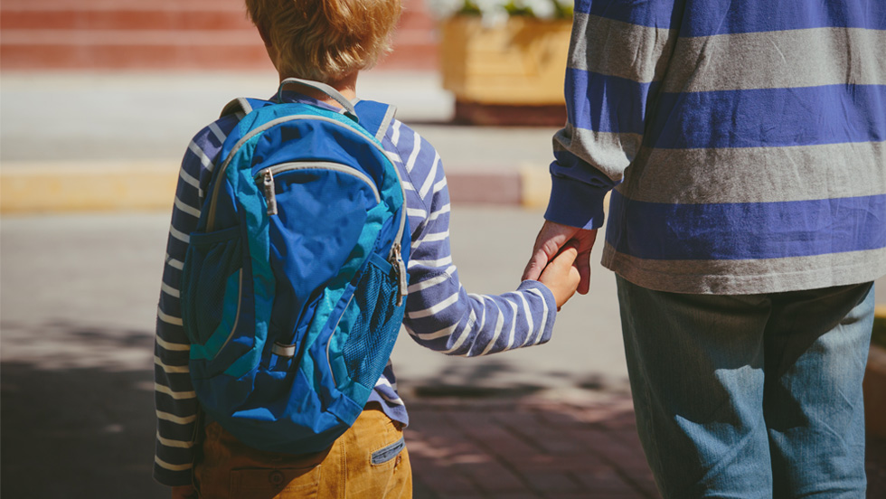 A young child, seen from behind, wearing a blue backpack and a blue and white striped long-sleeve shirt, holds hands with an adult, also seen from behind, wearing a blue and grey striped shirt and jeans. They are walking away from the camera, possibly towards a school building, which is faintly visible in the background.