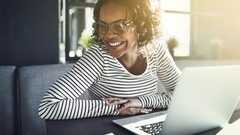 A happy, laughing Black woman wearing glasses and a horizontal black and white striped long-sleeve shirt is sitting at a table with a silver laptop in front of her. The setting appears to be a bright, sunny indoor space, possibly an office or cafe, with sunlight streaming in from the right. She has her hands clasped on the table and is looking down at the laptop screen.