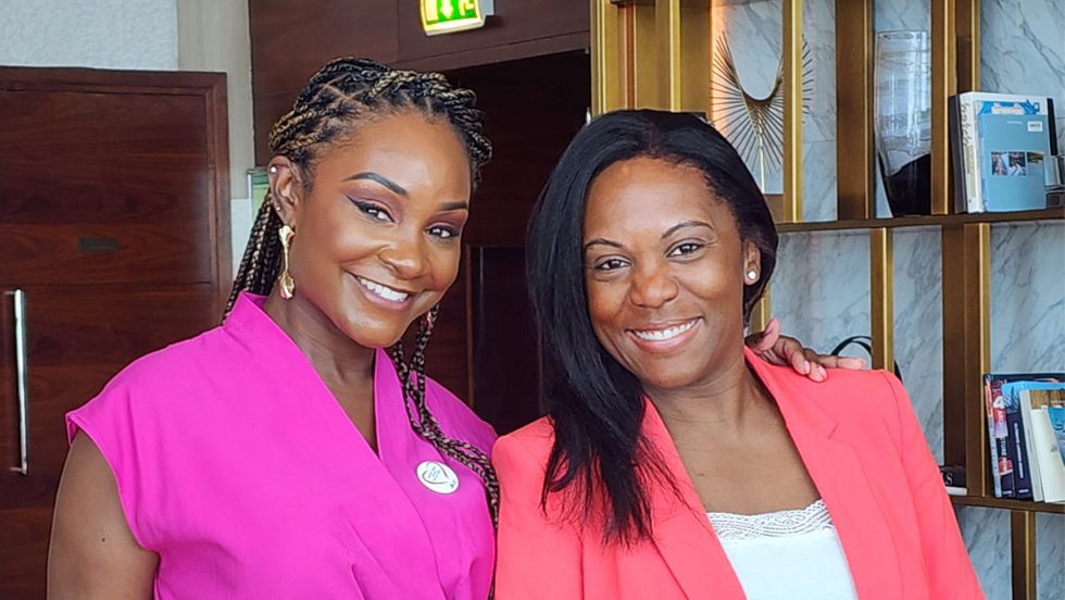Two women smiling and standing close together. The woman on the left is wearing a bright pink top and has braided hair, while the woman on the right is wearing a coral blazer over a white top.