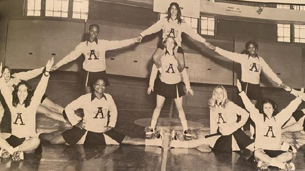 Adelphi cheerleaders in formation on the basketball court. Four cheerleaders are seated on the floor, five are behind them, with one cheerleader carrying another on her shoulders.