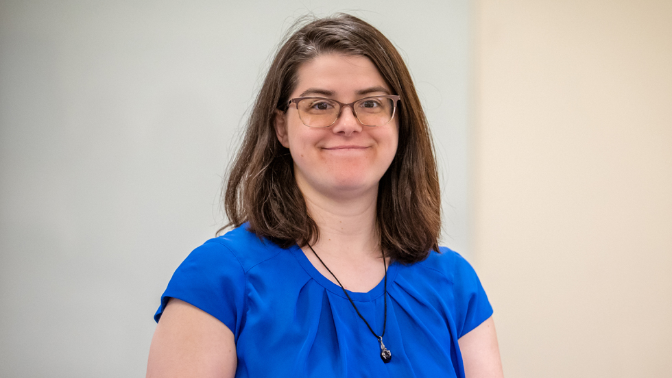 A woman with shoulder-length brown hair wearing glasses, a royal blue blouse, and a black necklace, smiling.