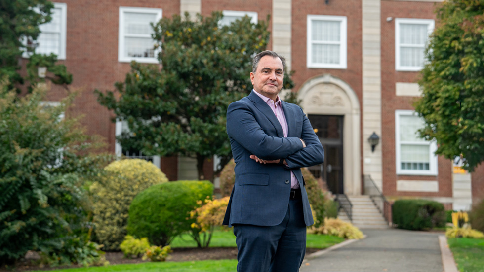 A man in a blue suit, looking confident, stands in front of a historic building on Adelphi's college campus.