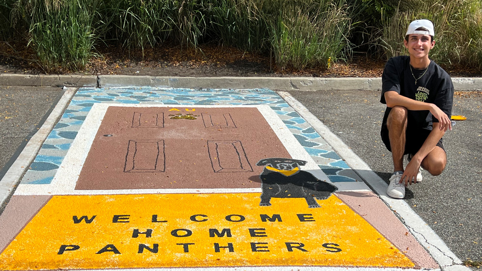A young man, James Tullo, kneels by his parking spot design, labeled "Welcome Home Panthers"
