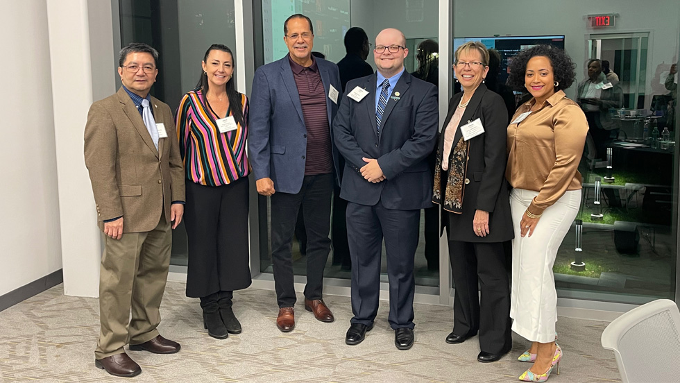 A group of six professionally dressed individuals, three men and three women, stand side by side indoors, posing for a photo. They are smiling and name tags are visible on each person. The setting features a modern office environment with glass walls, reflections and a conference room visible in the background.